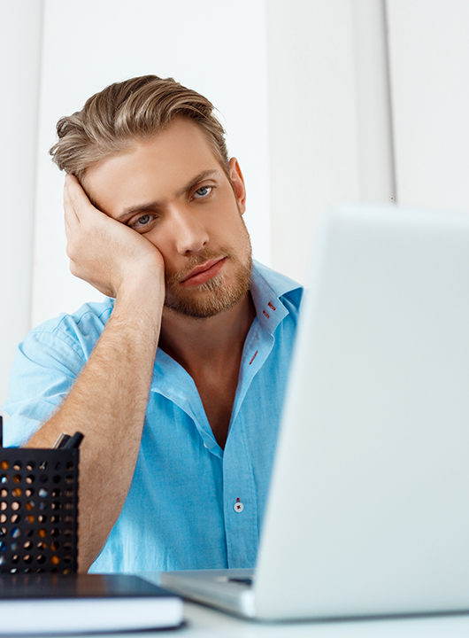 Young handsome tired confident pensive businessman sitting at table working on laptop with cup of coffee aside. White modern office interior background.